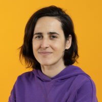 white non-binary person with brown hair siting in front of a yellow background wearing a purple sweatshirt