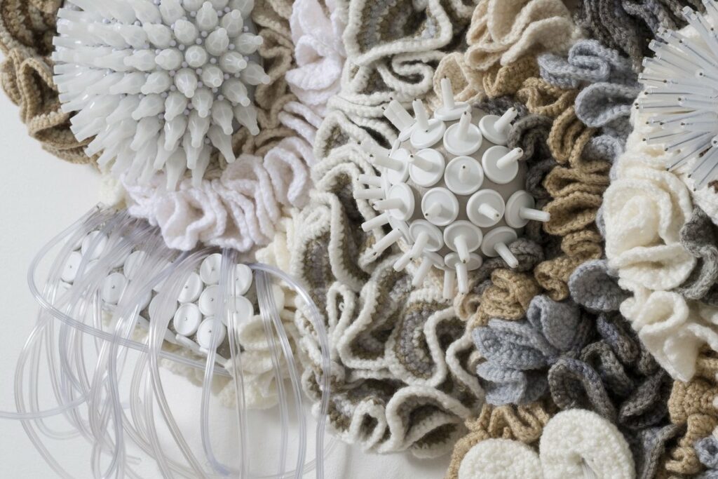 Image Description: A close up photo of a densely textured crochet wall sculpture in white, faded gray, brown and light browns. It is a tightly woven body of delicate wrinkles, ruffles, curls undulating around three rounded mounds. The mounds are covered with white or translucent spiked tips and short, stiff thin tubing. Long thin tubing stretches from its bottom edges in small patches.